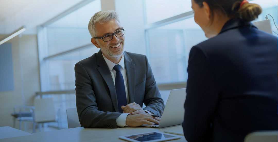 Two people in business attire sitting across from each other at a desk with a laptop and tablet.