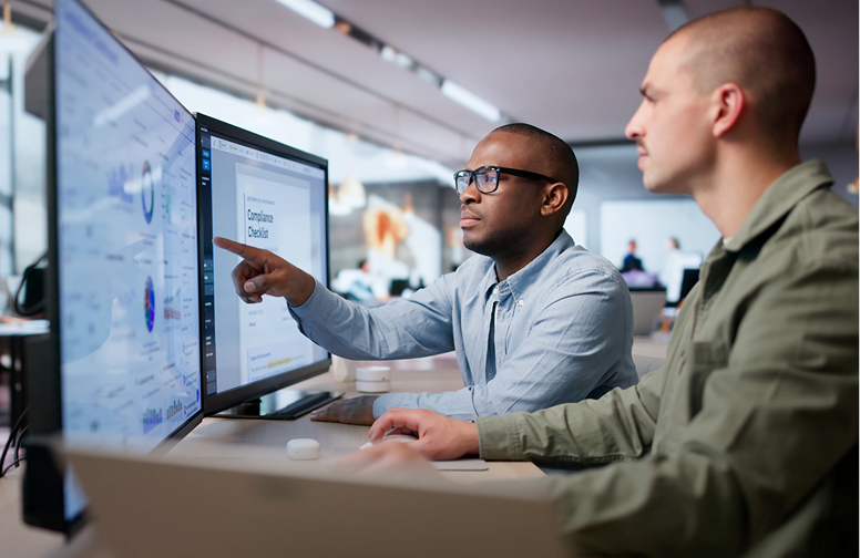Two men at desks looking at computer screens with charts and a compliance checklist.