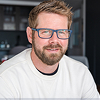 Man with short brown hair, beard, and glasses wearing a white shirt with blurred background.