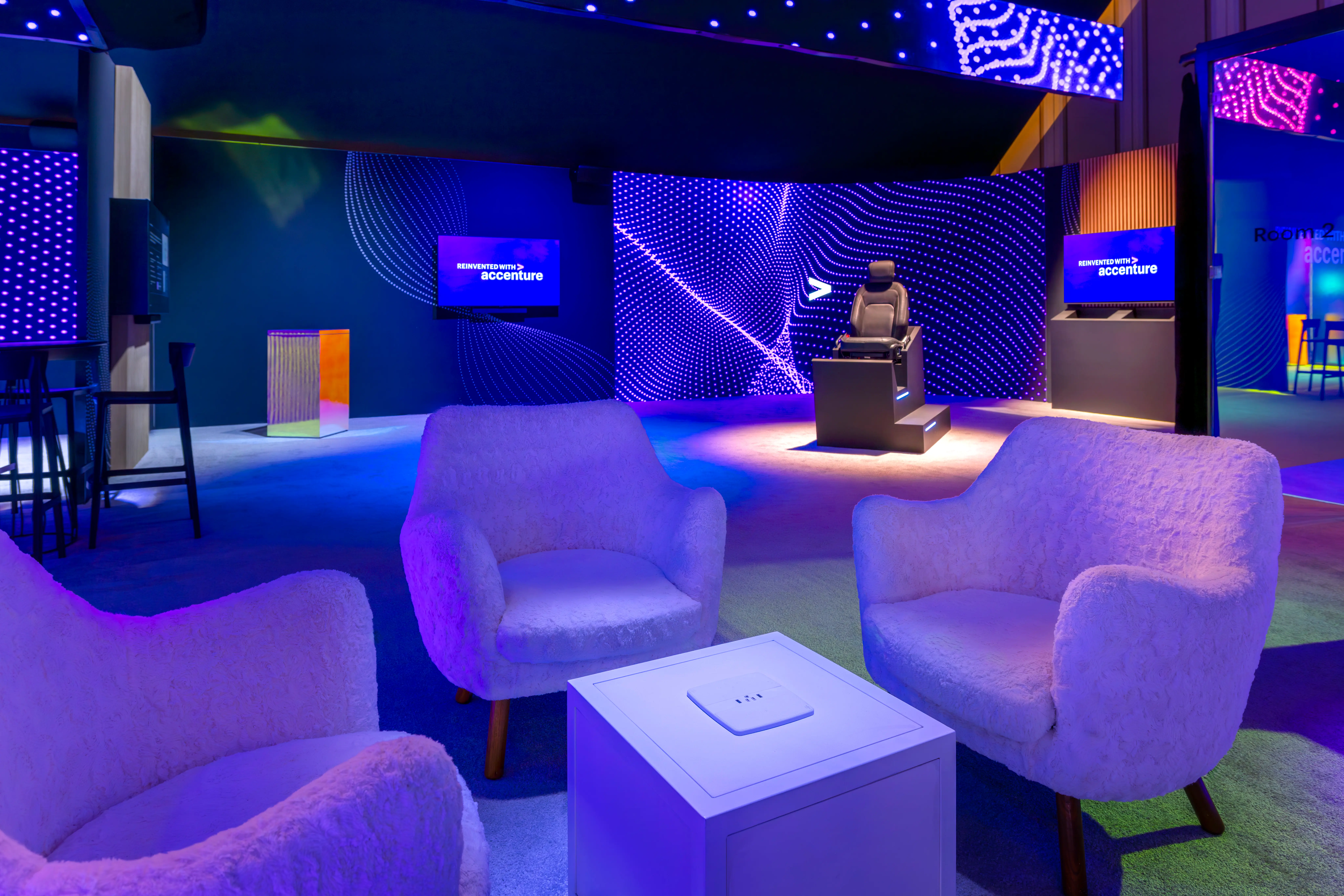 Modern lounge area with three white armchairs, small square table, and digital displays showing the Accenture logo in a room with blue and purple lighting and abstract wall designs.

