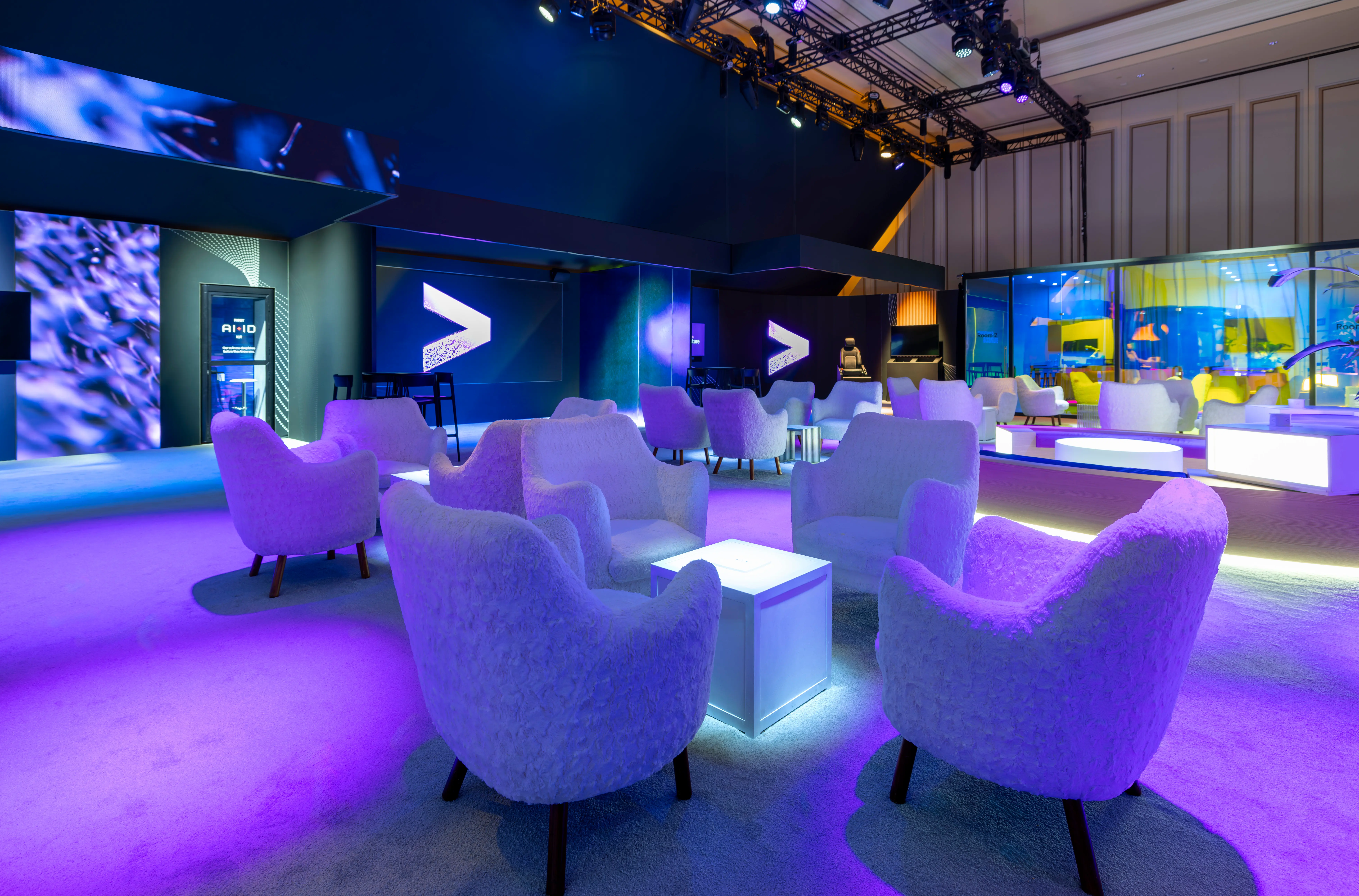 A modern lounge area with white armchairs and illuminated tables, bathed in purple and blue lighting, set in a spacious event venue.


