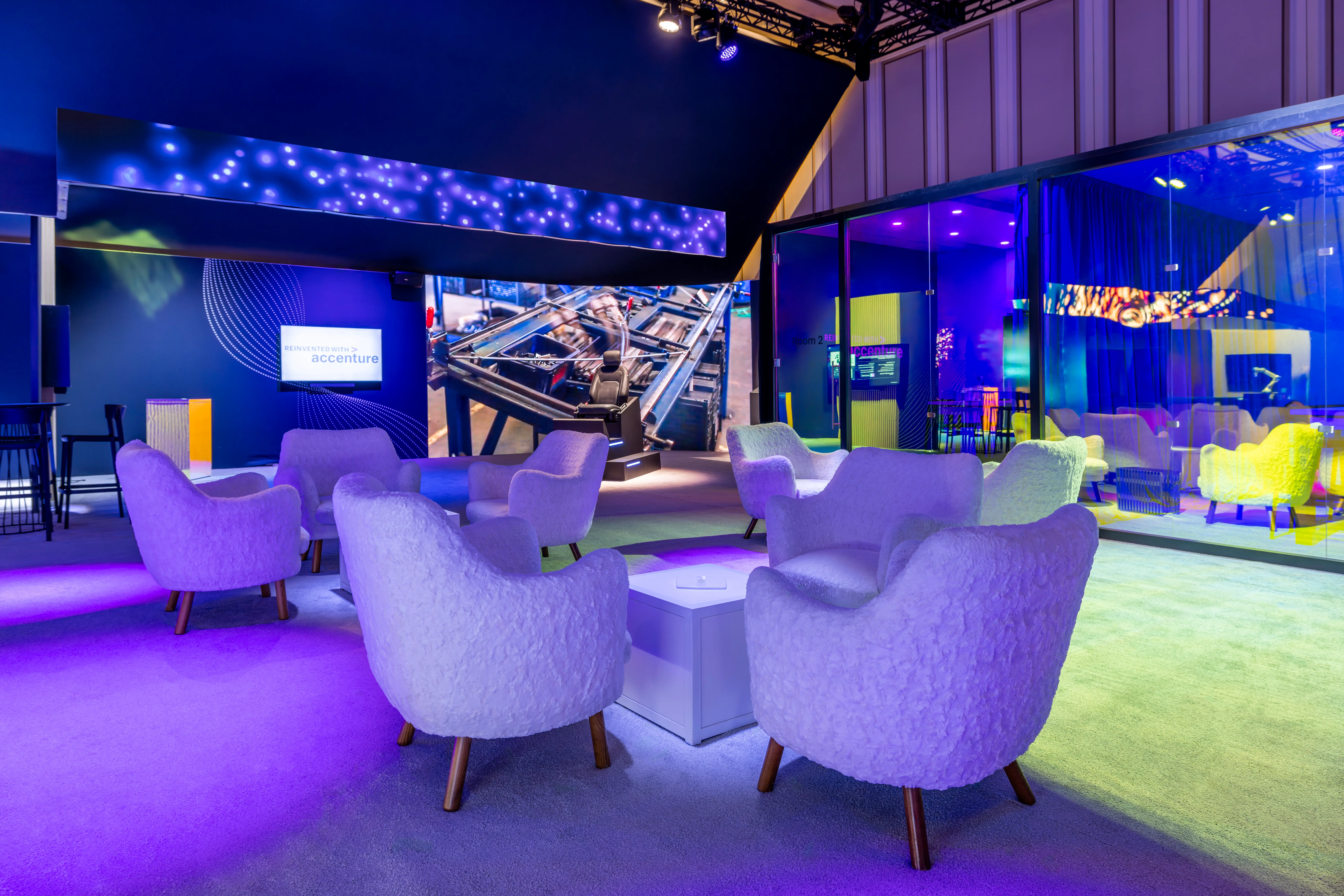 Modern lounge with plush, white chairs around a central table, illuminated by vibrant purple and blue lights. Screens display dynamic visuals, creating a futuristic, inviting atmosphere.