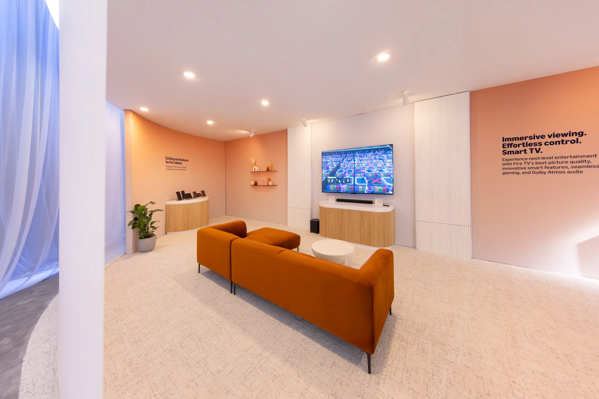 Modern living room with a sleek orange sofa facing a large TV on a wooden console. Soft lighting, light peach walls, and a potted plant add warmth.