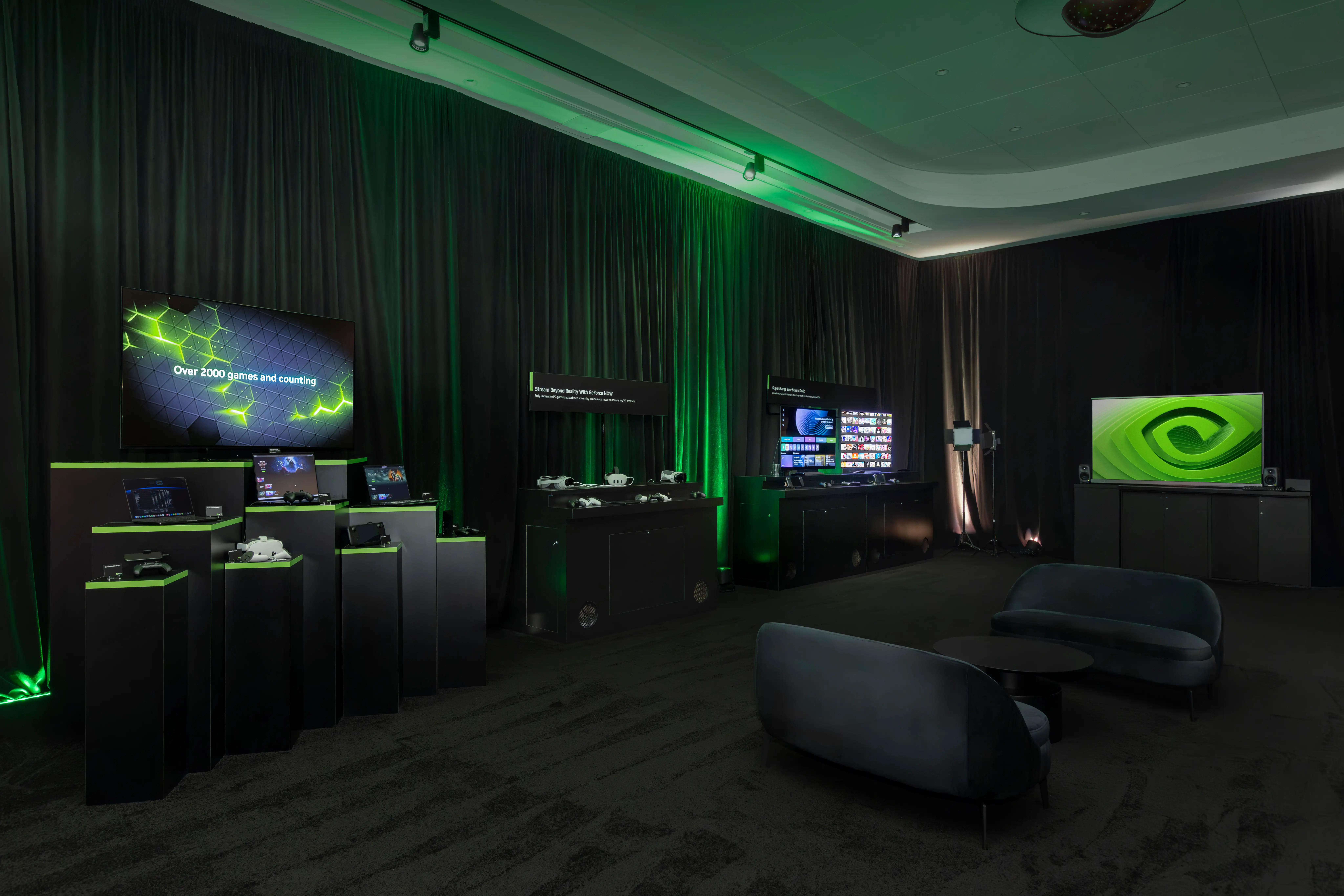 A sleek tech-themed room with green lighting features display screens showcasing gaming content. Modern black couches and round tables provide a futuristic vibe.