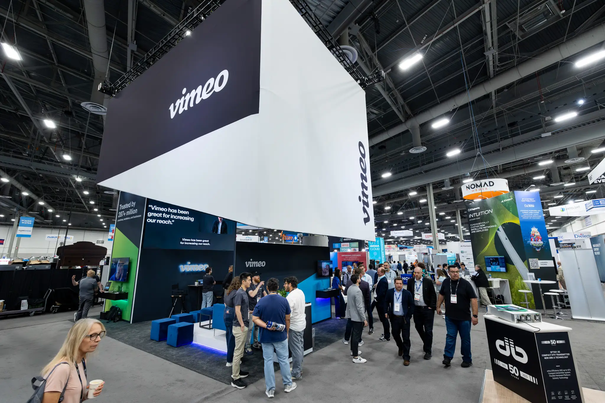 Spacious convention hall with a large Vimeo booth overhead and people interacting around it. Bright, modern design with a busy, engaging atmosphere.