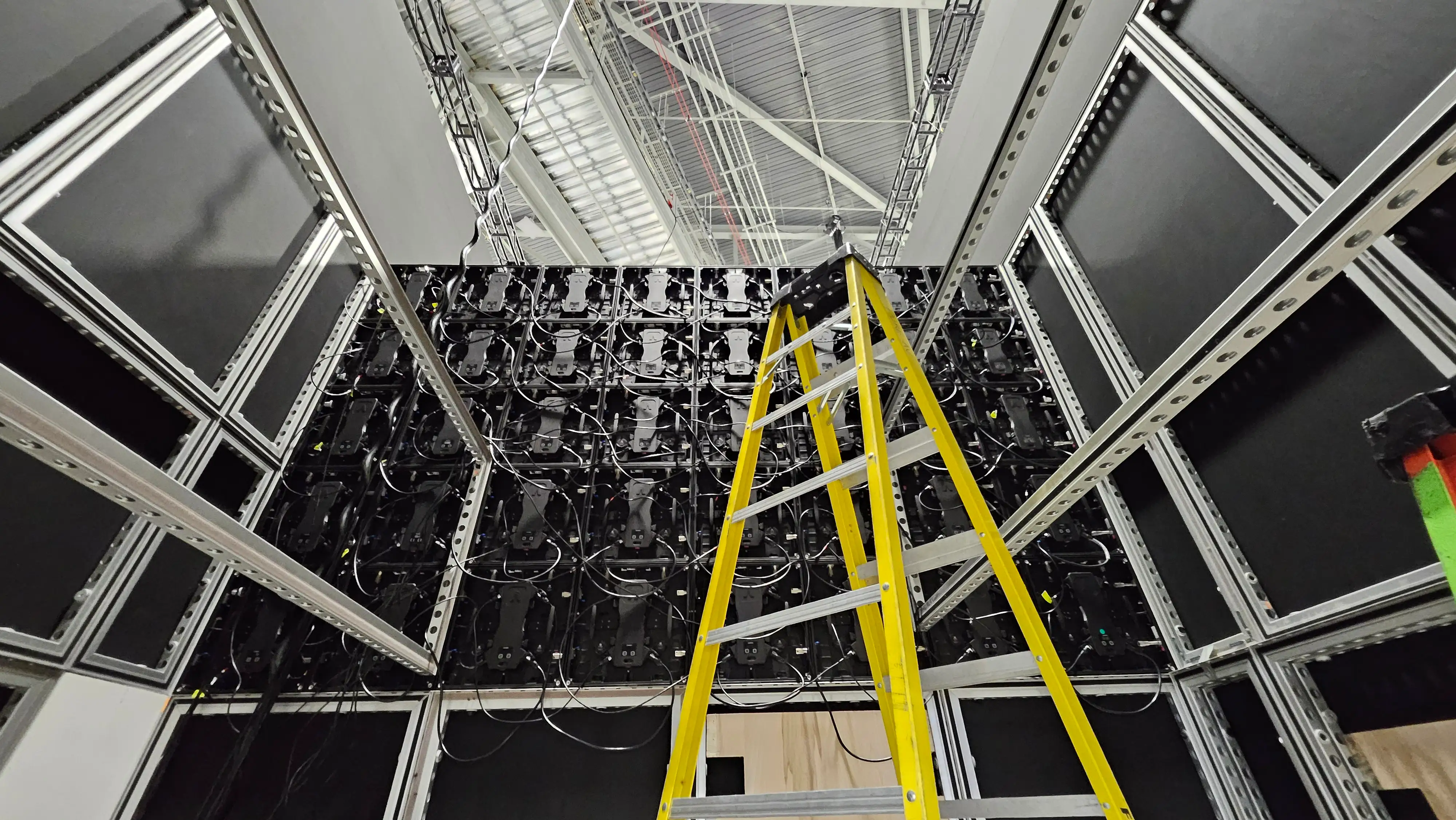 Yellow ladder in front of a large wall of interconnected LED panels and cables, under a high industrial ceiling. The scene is industrial and technical.