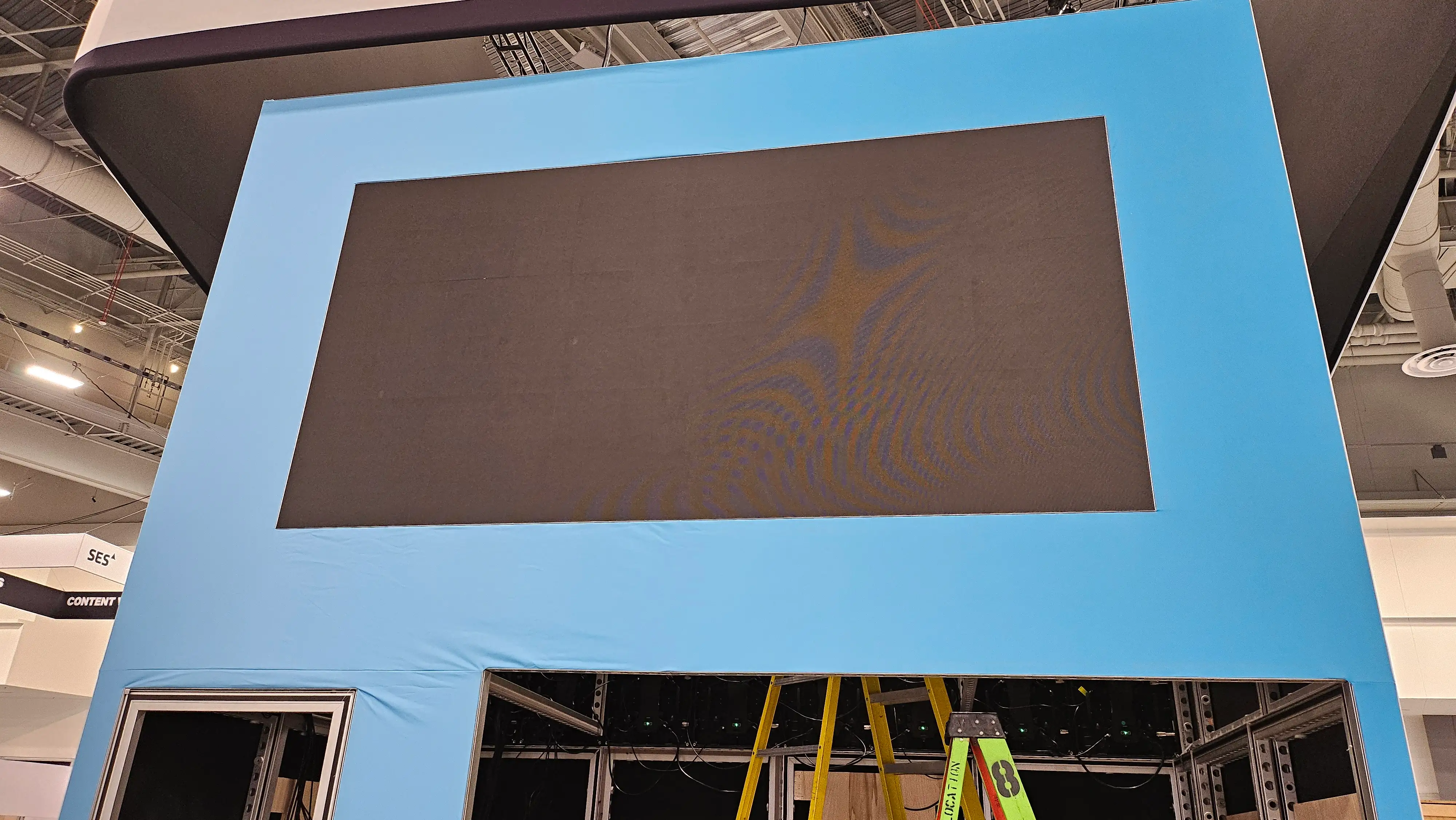 Large black screen framed by a blue structure, partially assembled at a trade show. Ladders and exposed wiring indicate ongoing construction. Bright, industrial setting.