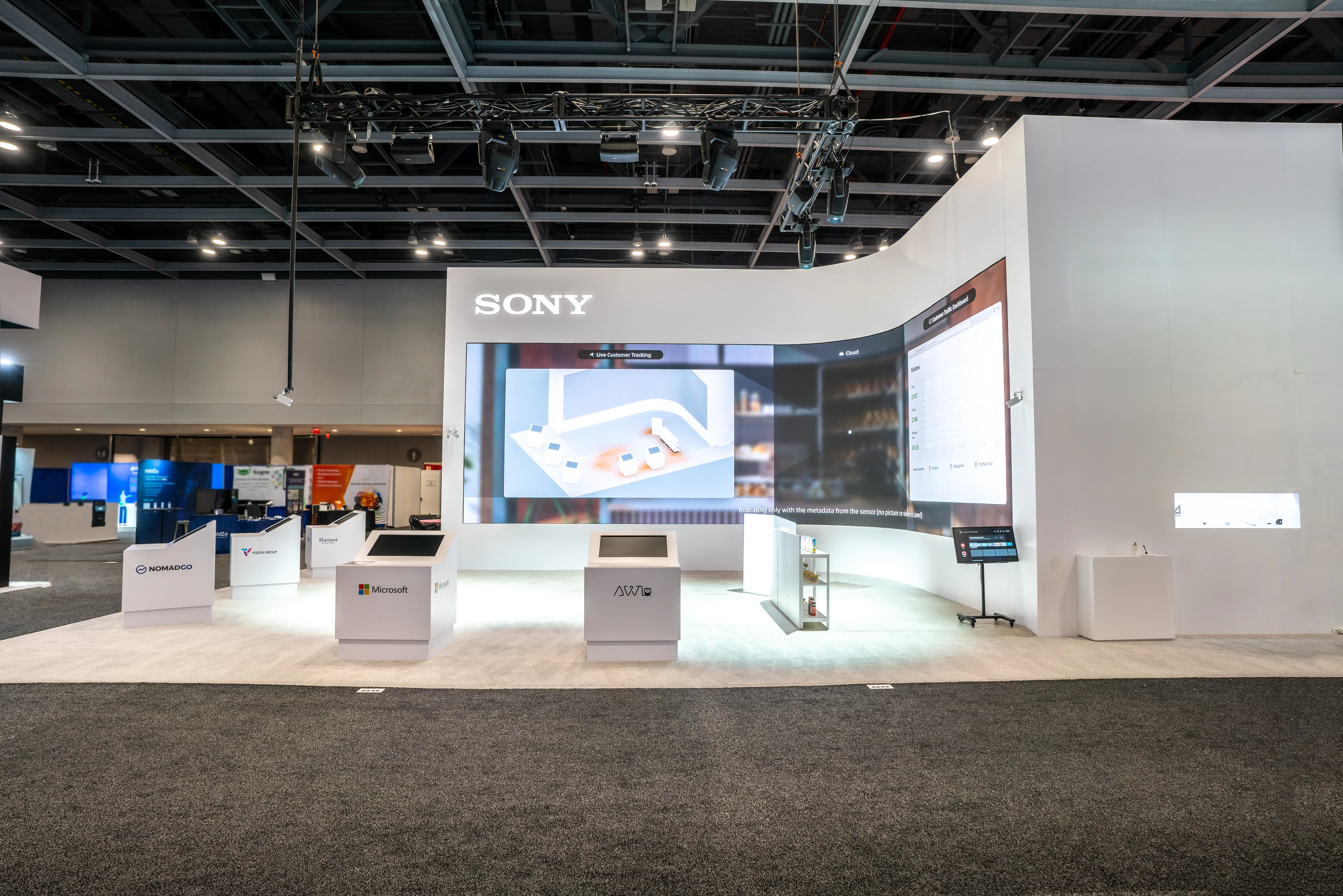 Spacious tech exhibit with a large Sony screen displaying a 3D model, surrounded by branded kiosks like Microsoft. The scene is modern and high-tech.