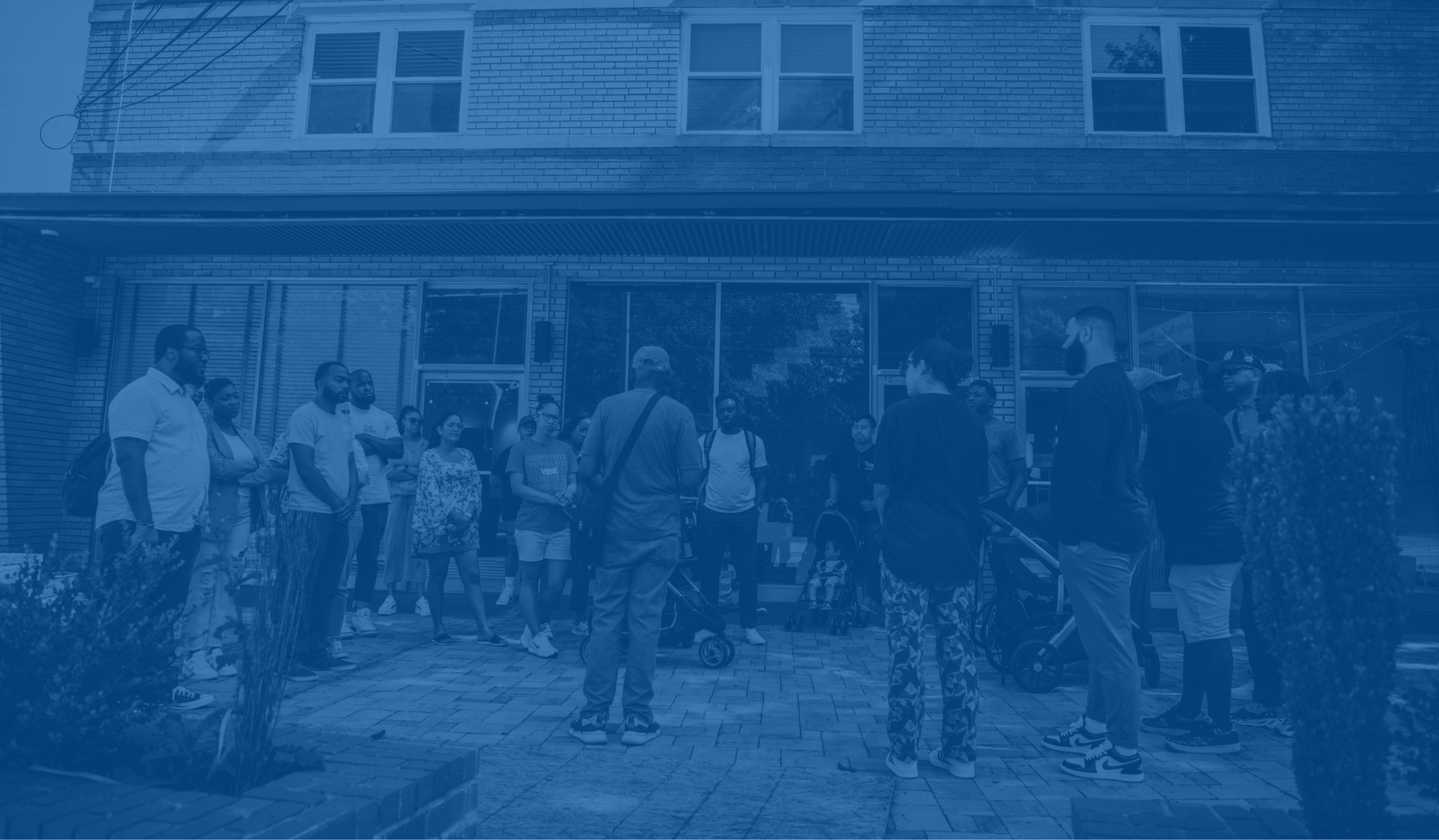 Group of diverse people gathered outside a brick building, some standing and others with strollers.