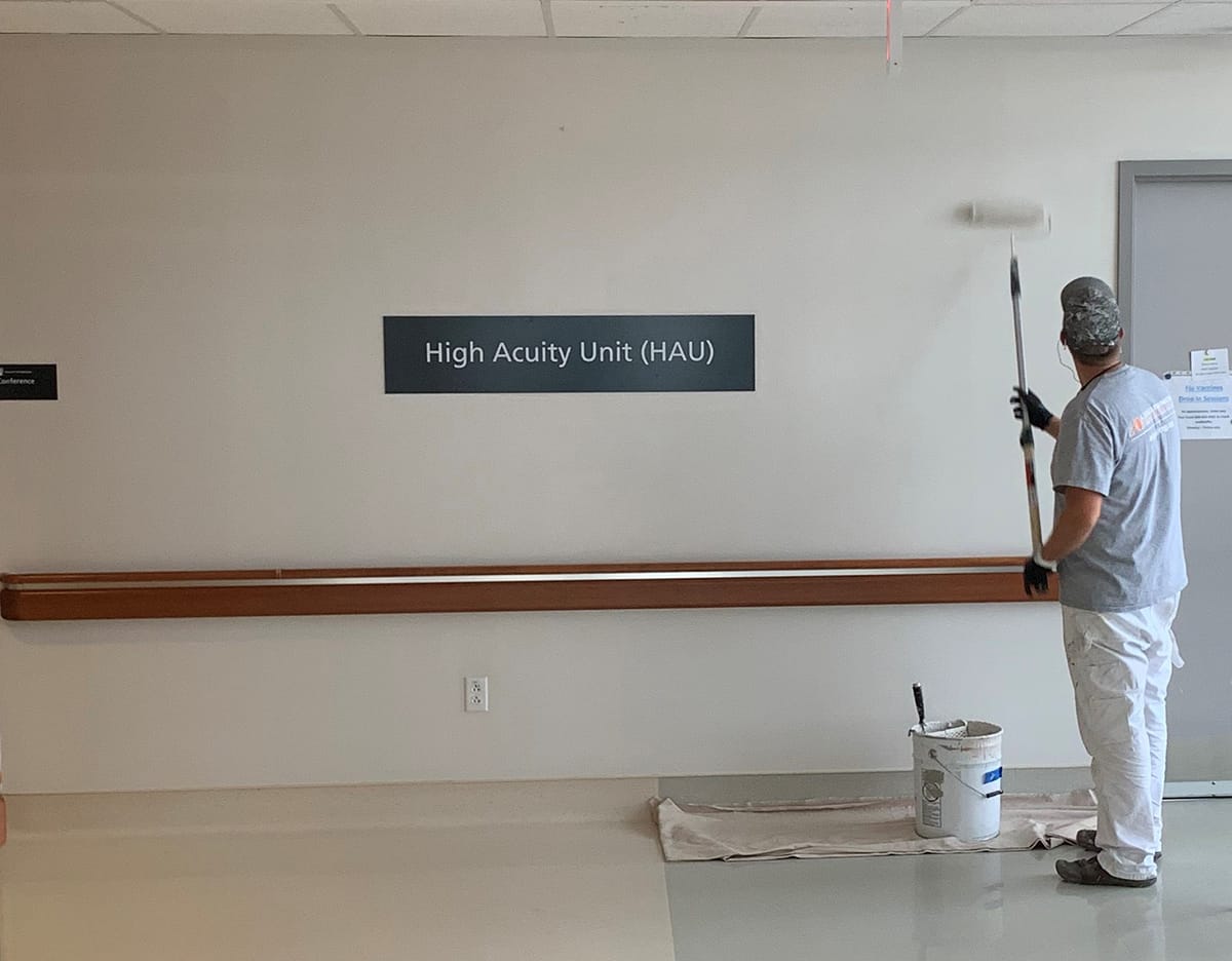 A worker in white pants and gray shirt painting a light beige wall next to a sign reading 'High Acuity Unit (HAU)' with a paint roller.