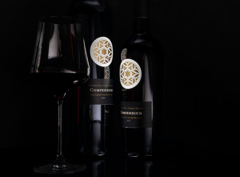 Two bottles of Compendium wine and a glass of red wine against a black background.
