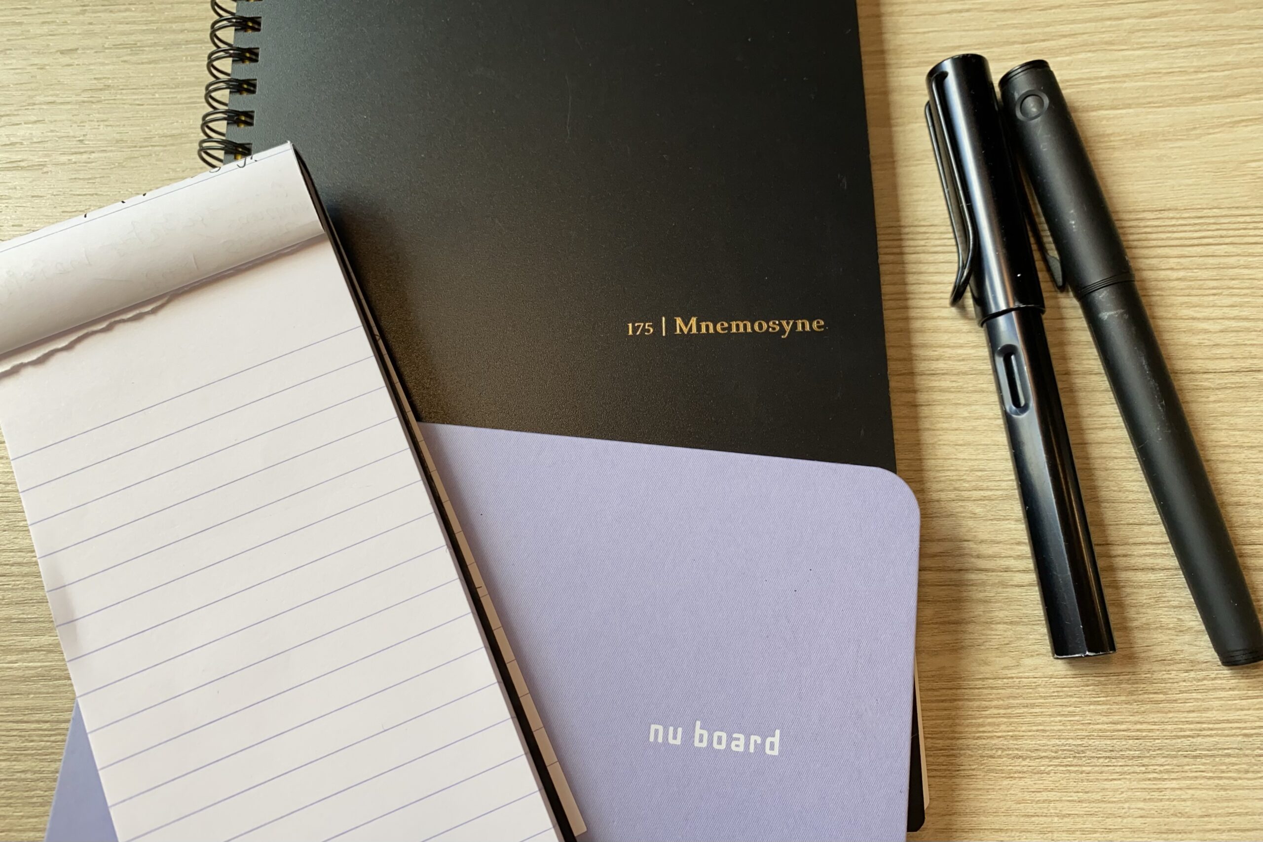 Desktop calendar, long skinny notepad, black notebook, purple notebook, two fountain pens and a sticky note pad on a desk.