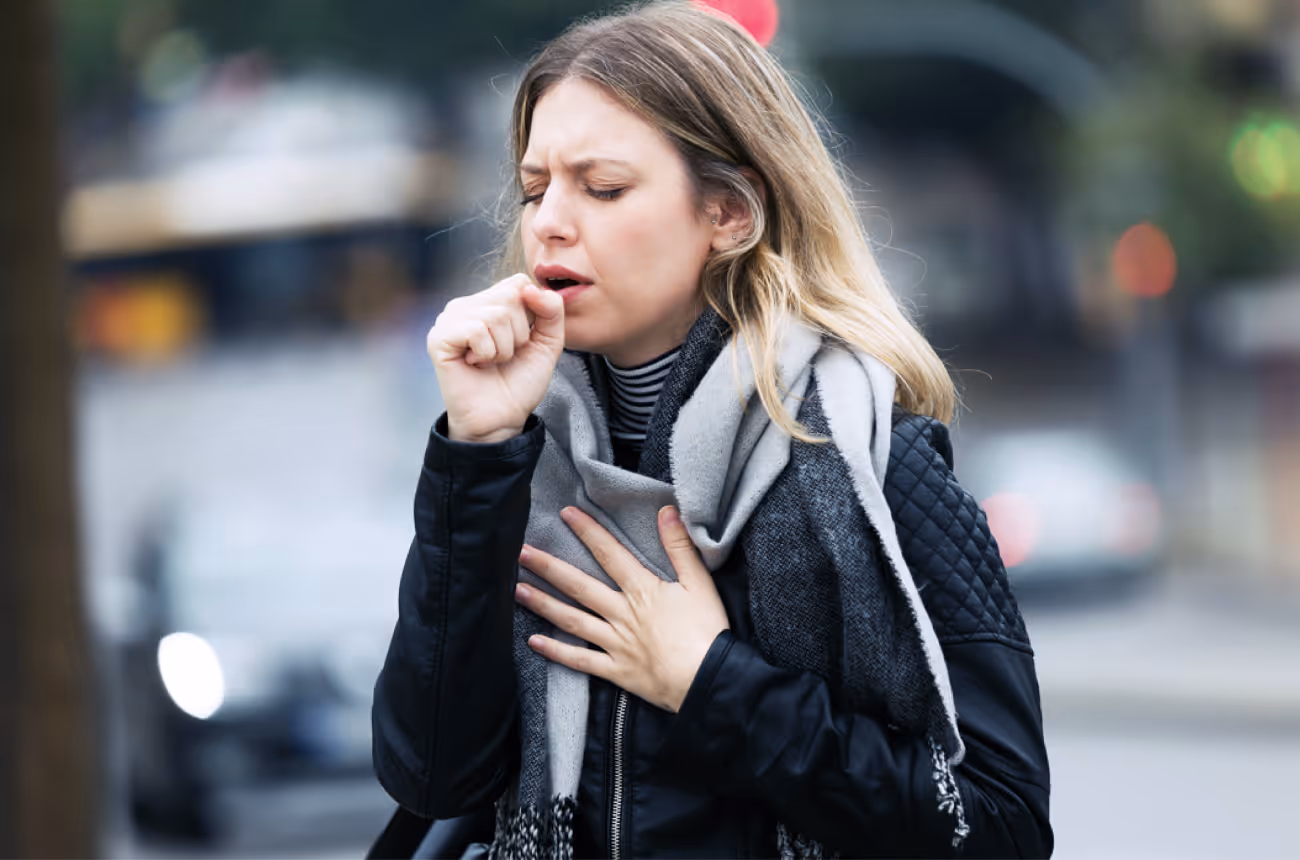 young blonde woman coughing into a closed fist while standing outside