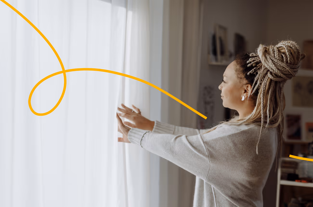 Woman with blonde hair in a bun opening curtains on a window