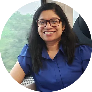 Woman with long dark hair and glasses smiling wearing blue blouse sitting by window with greenery.