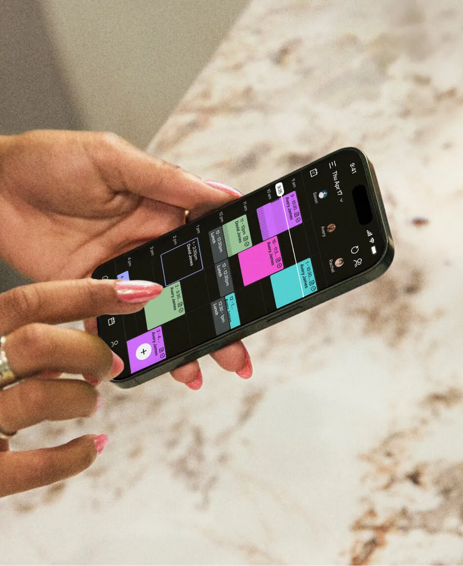 Manicured hands holding a smartphone displaying a color-coded calendar demonstrating beauty salon software.