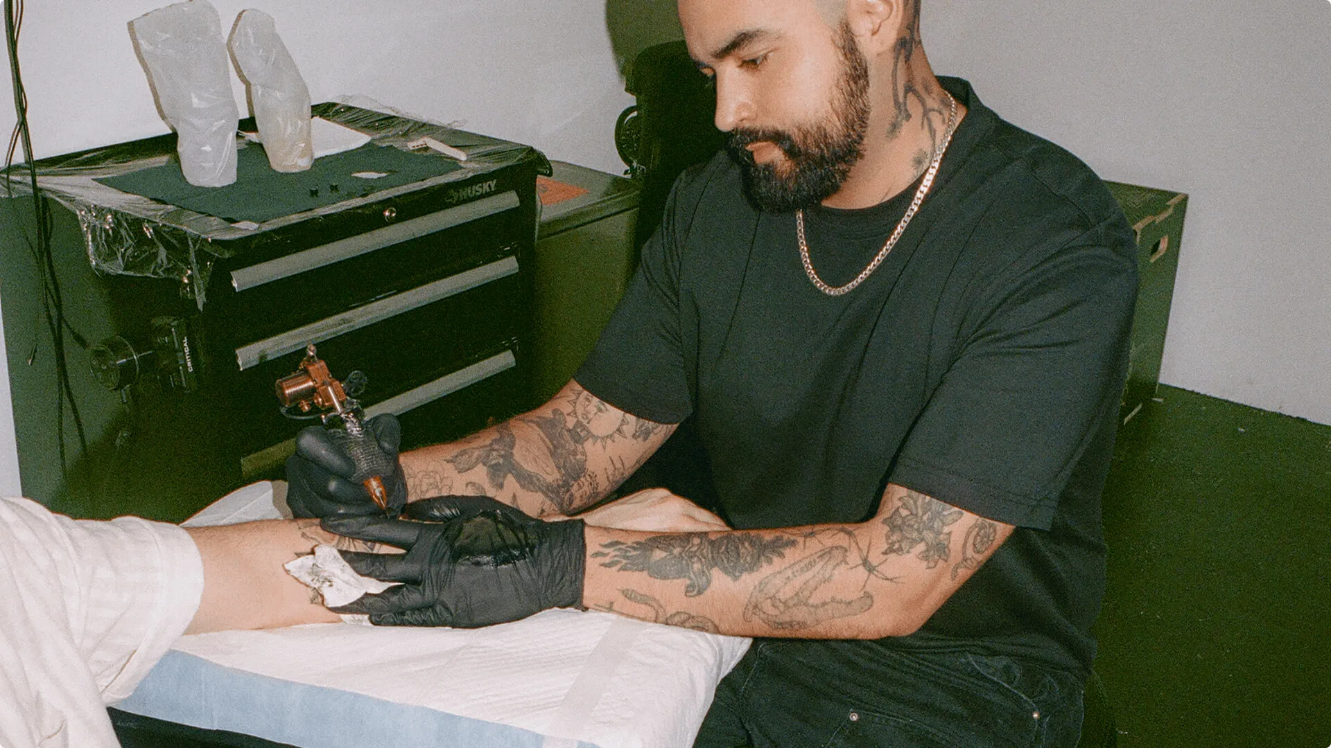 A male tattoo artist in a black t-shirt and gloves inking his client’s forearm 