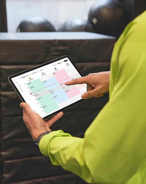 Hands holding a tablet displaying the GlossGenius calendar interface with color-coded appointments.
