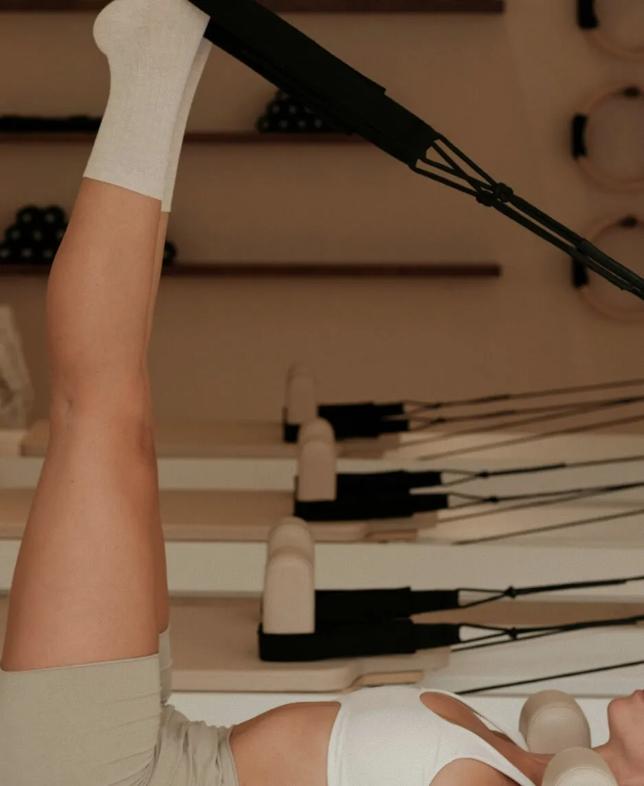 A girl in a neutral-colored workout outfit and pilates socks in a pike position on a pilates reformer.