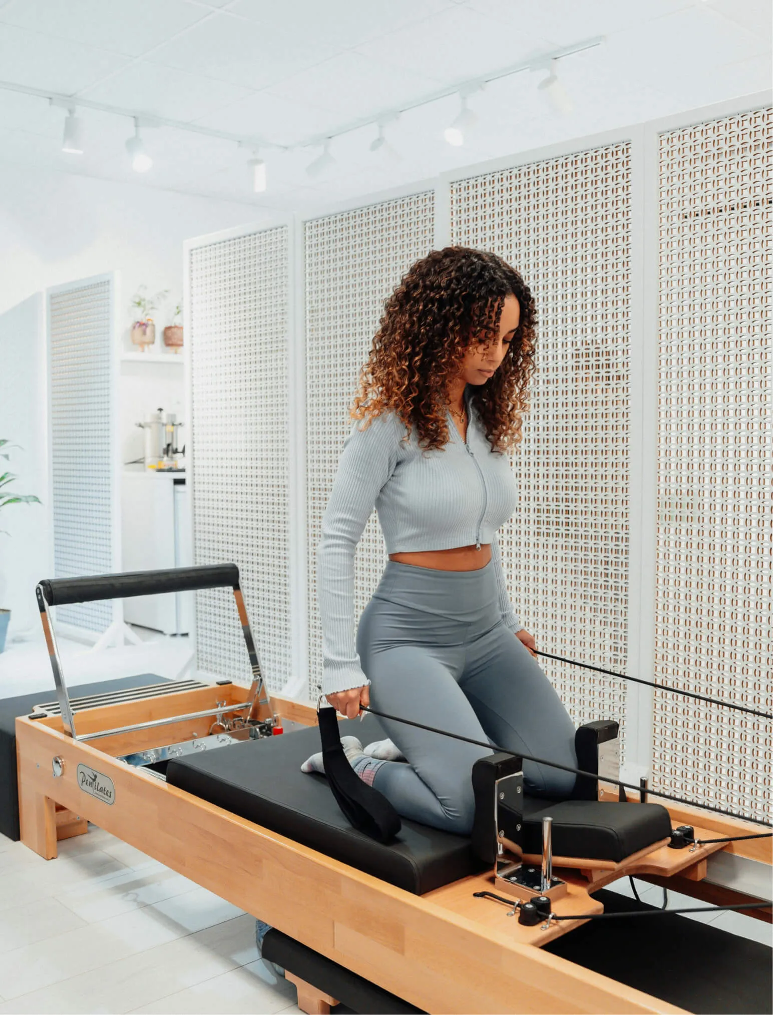 Woman with curly hair and a gray workout outfit on a reformer in a pilates studio.