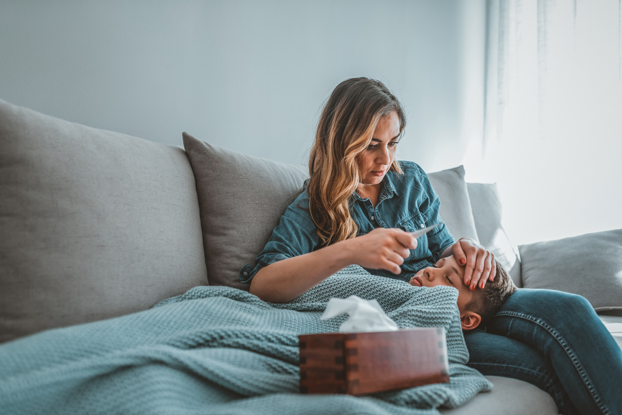 A little boy laying on the couch, on his mothers lap with a blanket on him and a box of tissues nearby. The mom is holding a thermometer, reviewing his temperature.