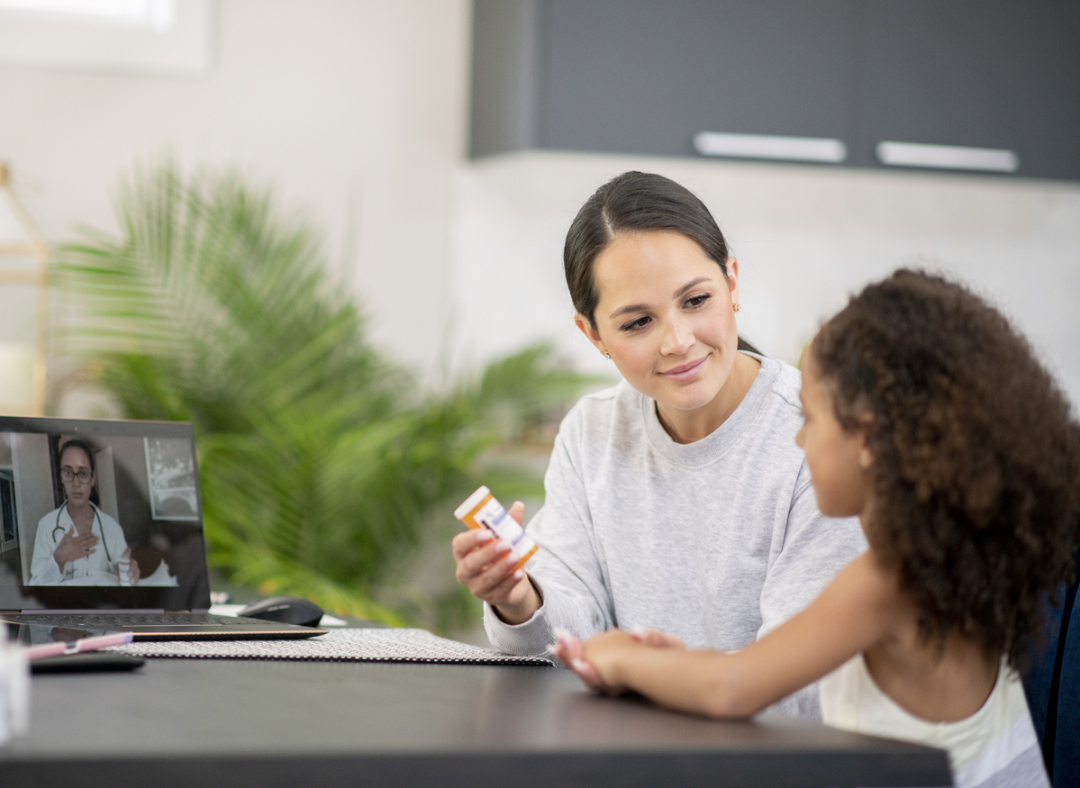 A mom holding a prescription medication bottle, sitting next to her daughter. Both are on a virtual visit with a doctor on a laptop.