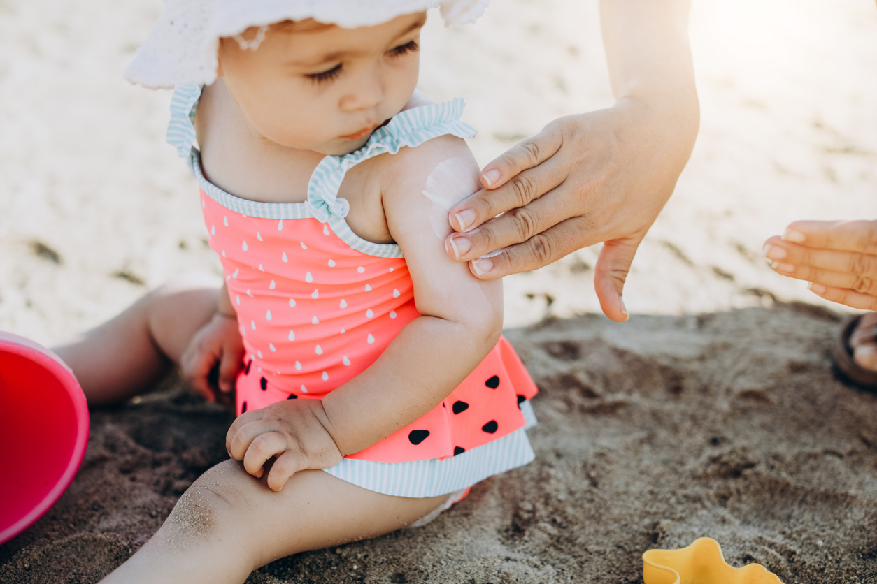 A baby girl sits on the sand at the beach in a watermelon swimsuit, watching as her parent applies suncreen on her arm.