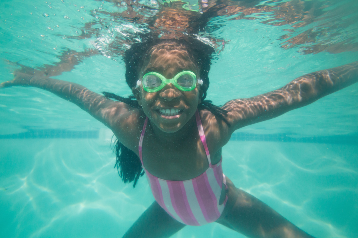 A little girl with braids, a pink swimsuit and green googles swims underwater in a pool.