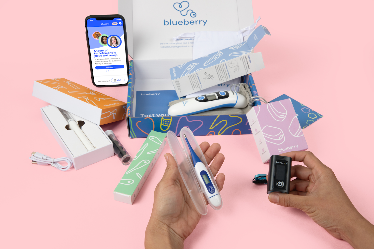 A person's left hand holding a thermometer and right hand holding a pulse oximeter. In the background, is a phone showing the Blueberry Pediatrics website, and a box with forehead thermometer.