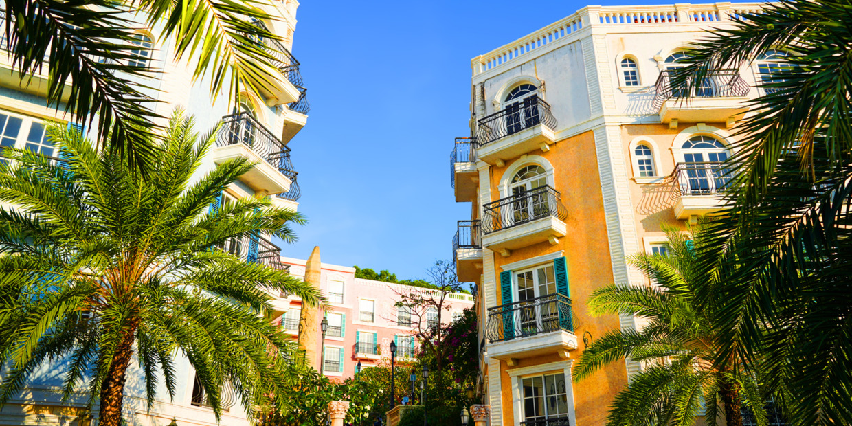 Palm trees surrounding colorful Mediterranean-style condominium buildings with balconies under a bright blue sky.