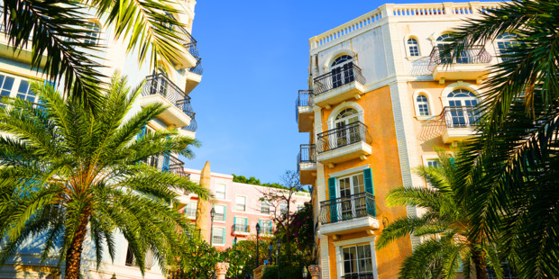 Palm trees surrounding colorful Mediterranean-style condominium buildings with balconies under a bright blue sky.