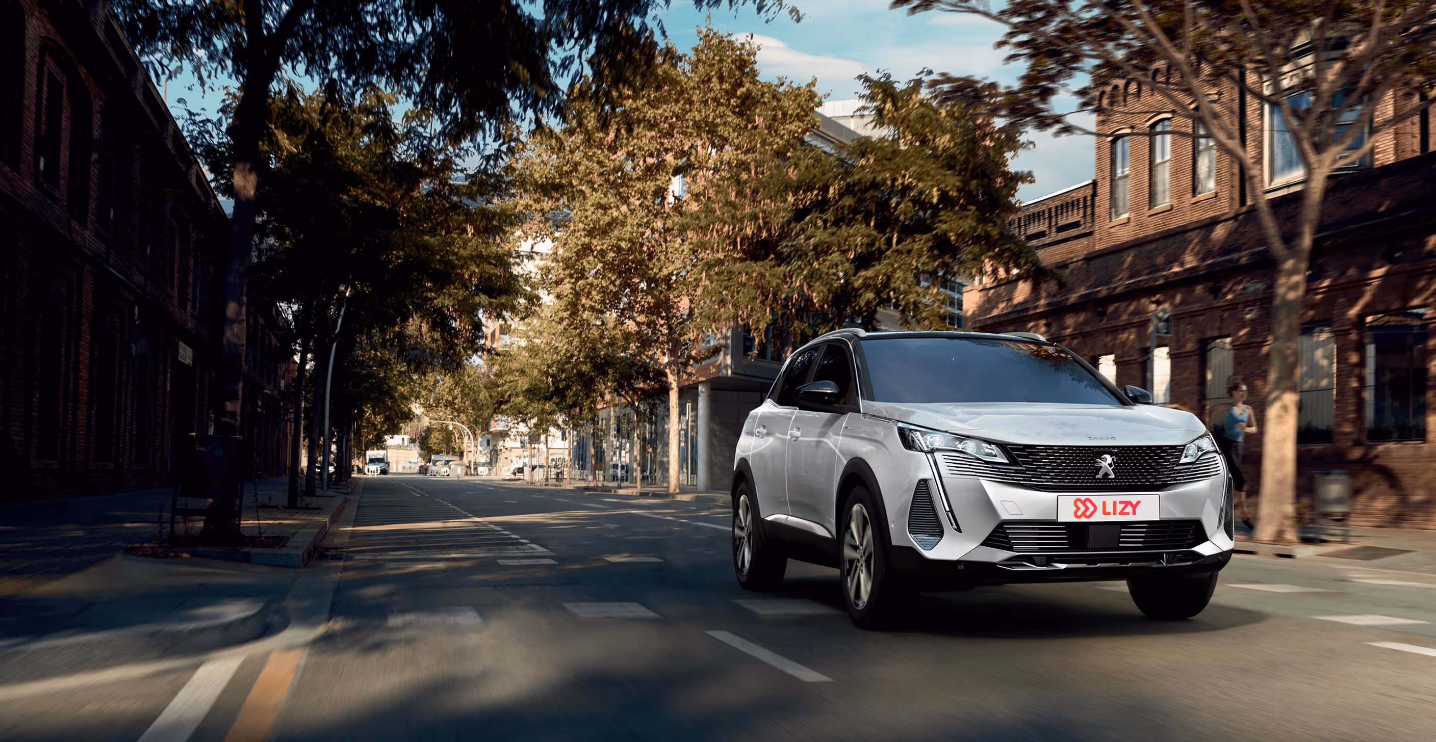 Silver Peugeot SUV driving down a tree-lined street with brick buildings and a pedestrian in the background.