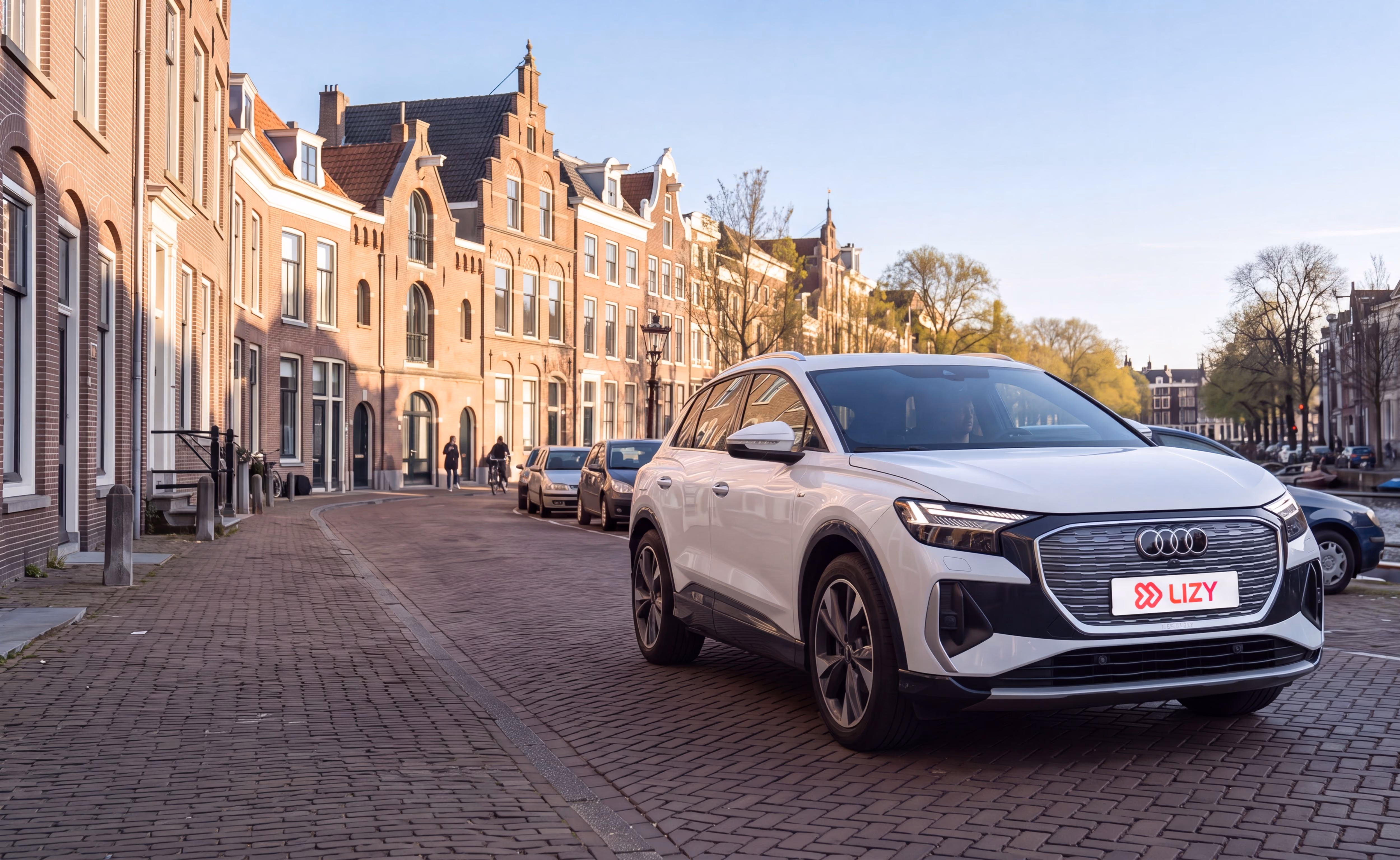 Witte Audi SUV Q4 e-tron geparkeerd op een geplaveide straat met historische bakstenen huizen en bomen.