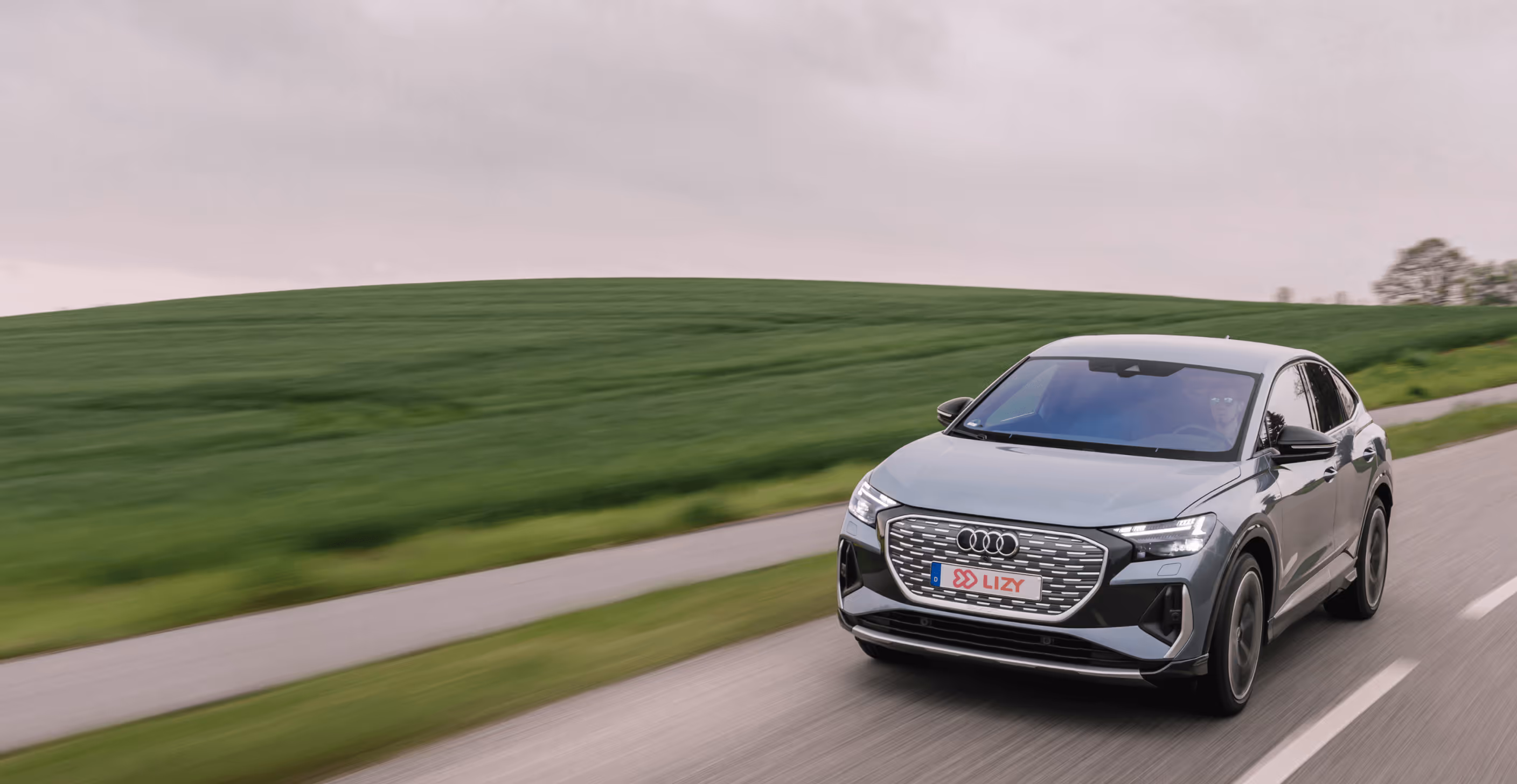 Silver Audi SUV driving on a country road with green fields and cloudy sky.