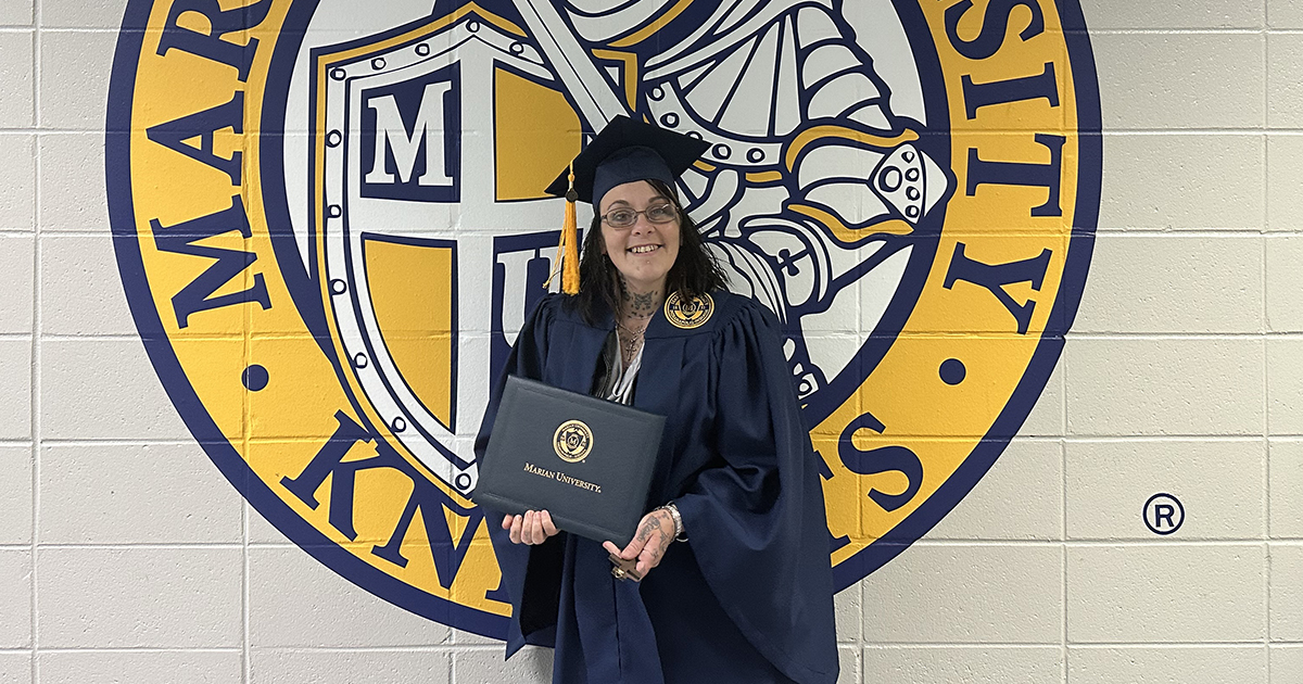 A graduate of WCP stands against a wall with the Marian University seal painted on it. She wears a graduation cap and gown, holding a diploma.