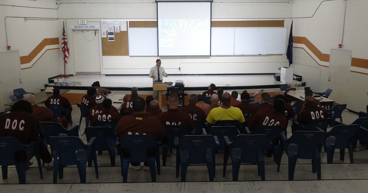 In a large room with tall ceiling a teacher stands facing a classroom in front of a projector screen