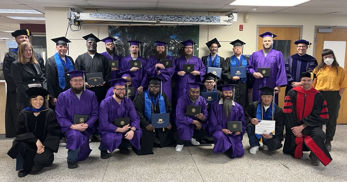 more than a dozen people pose standing in a group wearing cap and gown and holding diplomas