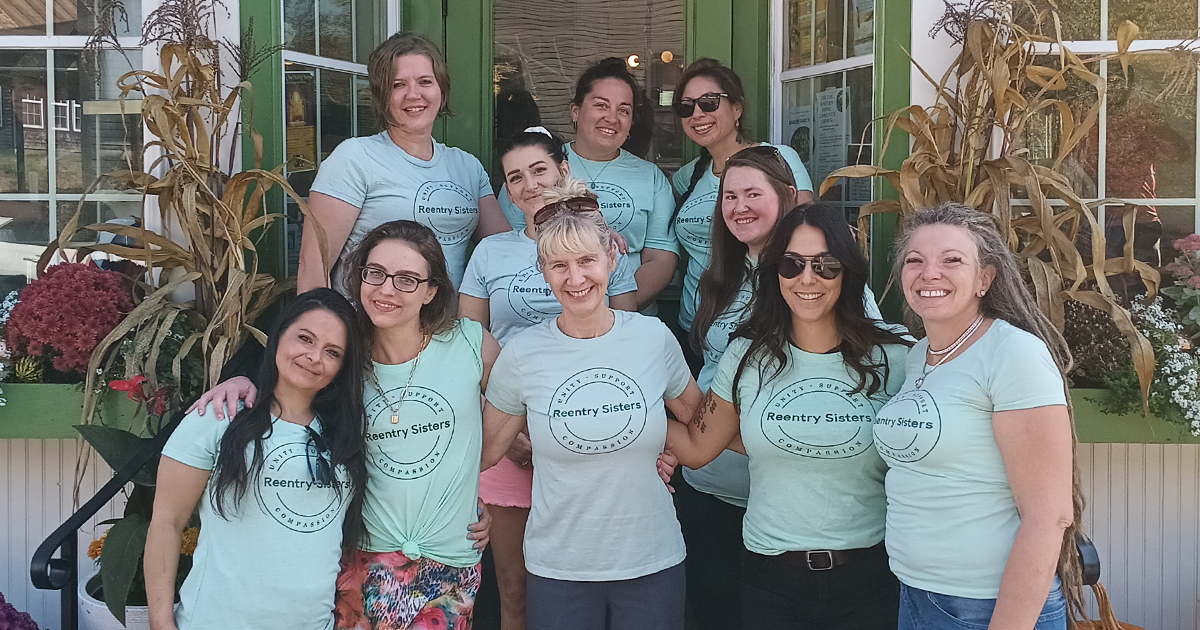 ten women wearing Reentry Sisters tshirts stand in front of a green building with fall decorations