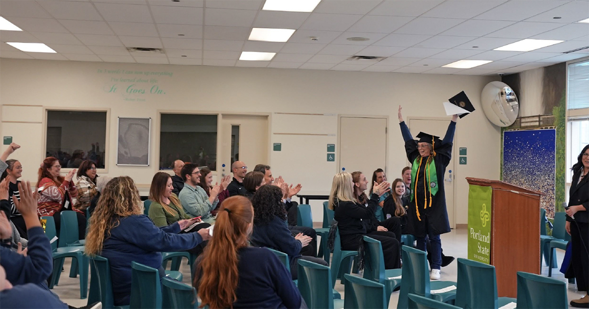 a snapshot of a graduate cheering at the front of a room with chairs arranged into rows for a small audience