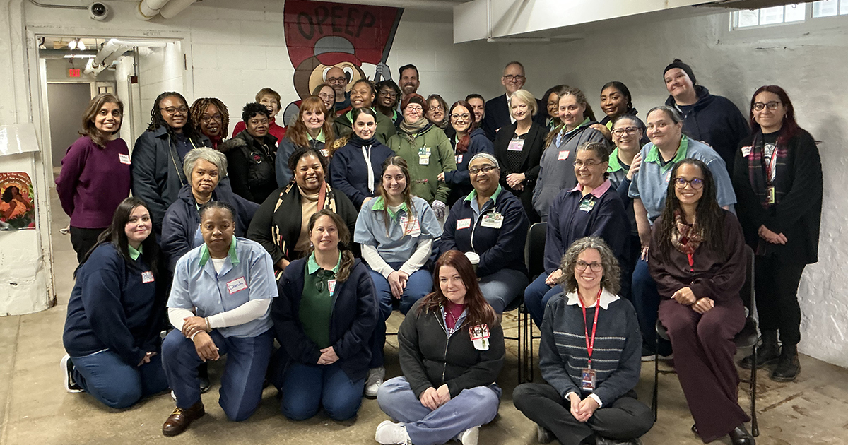 a group of about 30 people in casual professional attire pose together in a room with low ceiling and cement walls painted white with a OPEEP logo hand painted in dark red