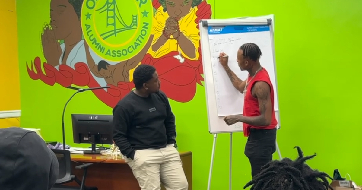 a man stands at an easel writing onto large paper while another man leans against a desk watching and others look on from the foreground. A bright green wall in the background with OAA logo painted on it.