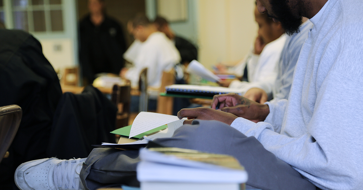 a closeup of a student sitting and studying with a white sweatshirt, jeans, white sneaker. Teacher and other students are blurred in the background. No faces visible.