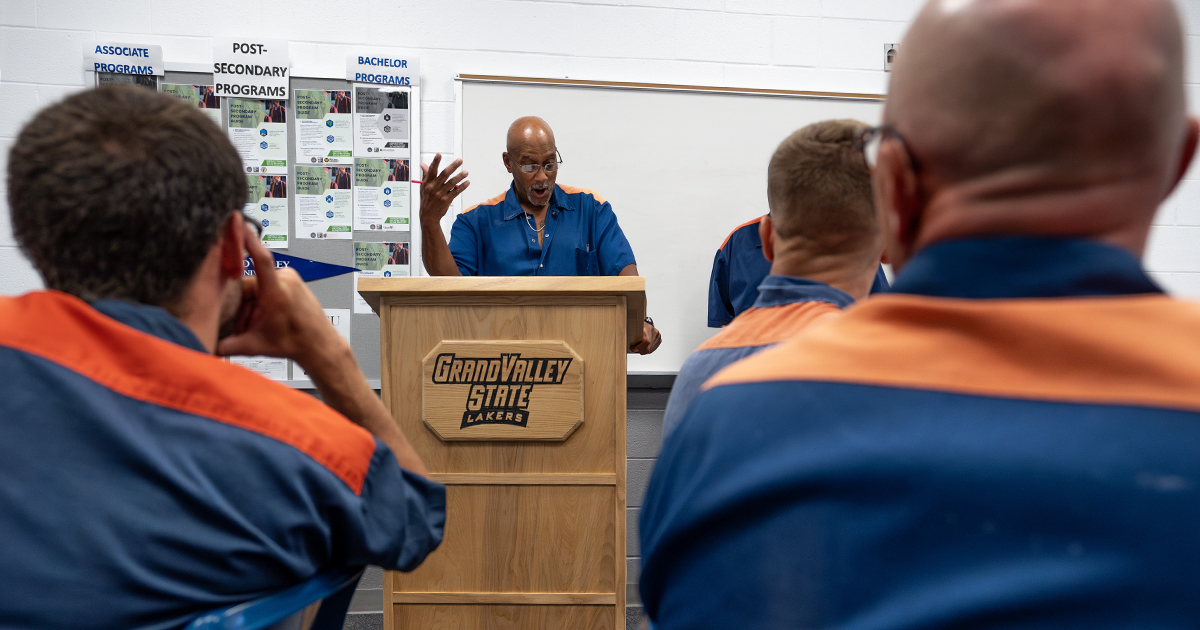 a man stands at a wooden podium gesturing and speaking. The podium has a plaque on the front with a logo "Grand Valley State Lakers" and in the background a whiteboard and a white brick wall with a bulletin board "Associate Programs, Bachelor Programs, Postsecondary Programs" with additional information on papers. An audience of other men all wearing the same blue and orange shirts is seen in the foreground (faces not visible).