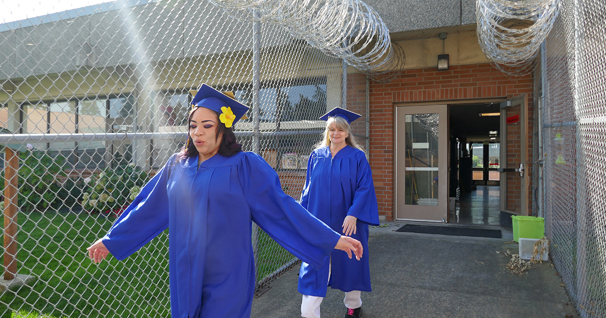 a woman in blue graduation cap and gown walks outside with arms outstretched with a joyful expression, a beam of light appearing before her. Another graduate walks behind her, both on a sidewalk lined by chain link and barbed wire.