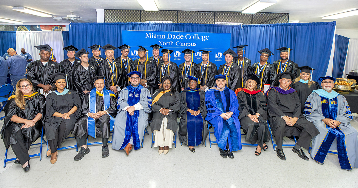 More than a dozen graduates pose together in a formal seated arrangement wearing shiny blue gowns in front of a shiny blue fabric backdrop