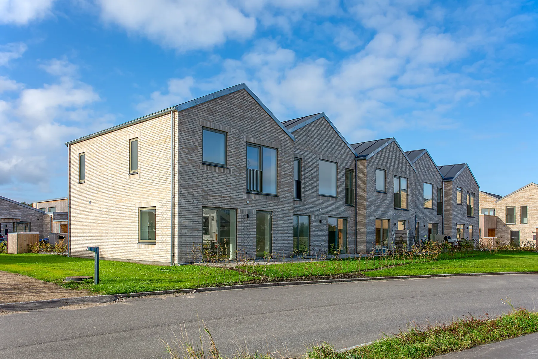 Modern row of light brick townhouses with large windows under a blue sky with scattered clouds.
