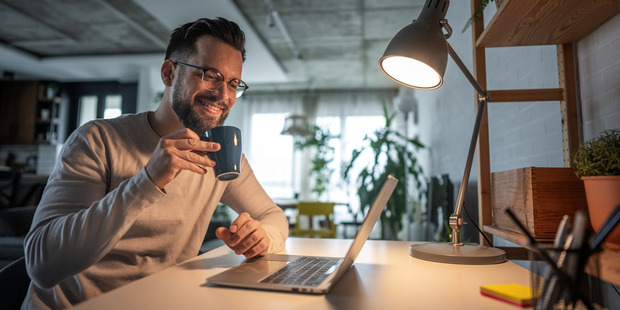 Man smiling at a laptop while holding a coffee mug, seated at a desk in a cozy home office with a lit desk lamp and plants in the background