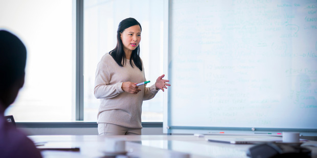 Woman presenting in a conference room while holding a marker and speaking beside a whiteboard filled with notes and diagrams.