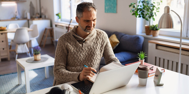 Man working from home at a desk, writing in a notebook beside a laptop and coffee.