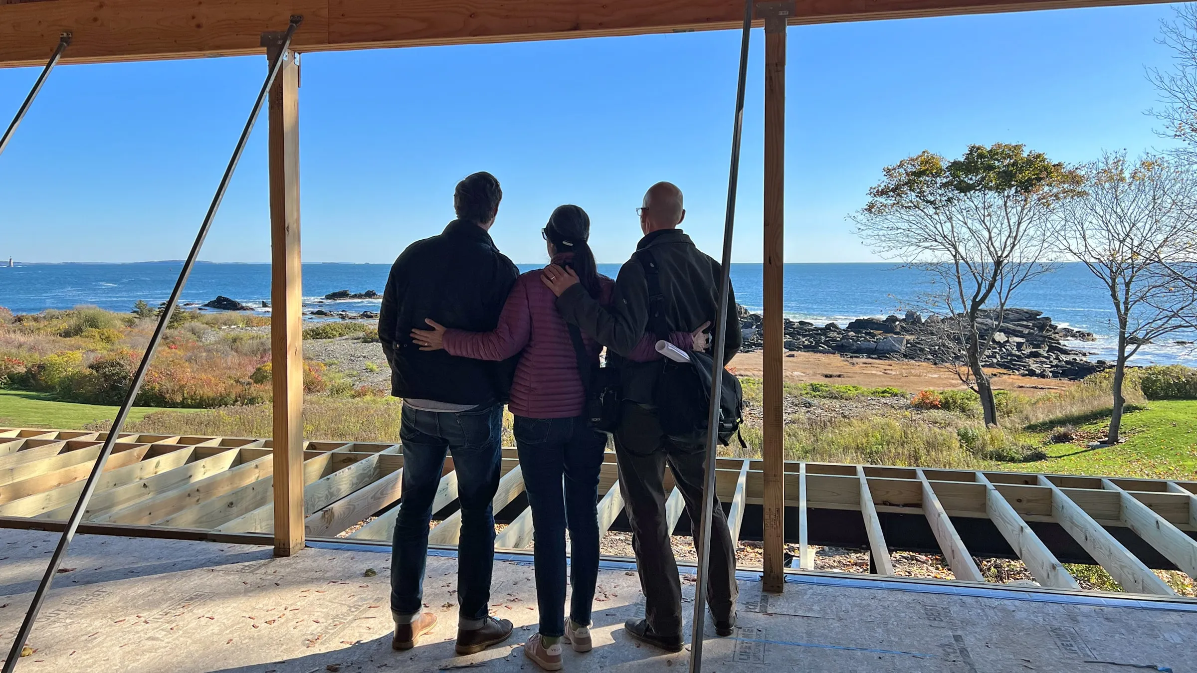Three people with arms around each other looking at the ocean from a partially built wooden deck.